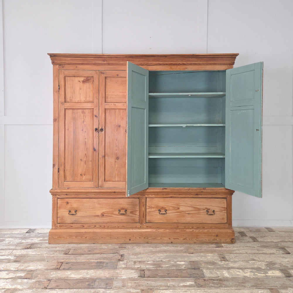 An antique Victorian pine housekeeper’s cupboard dating from the 19th century, with twin panelled doors to the upper section, fixed internal shelving, two base drawers with brass handles, and a simple plinth base.