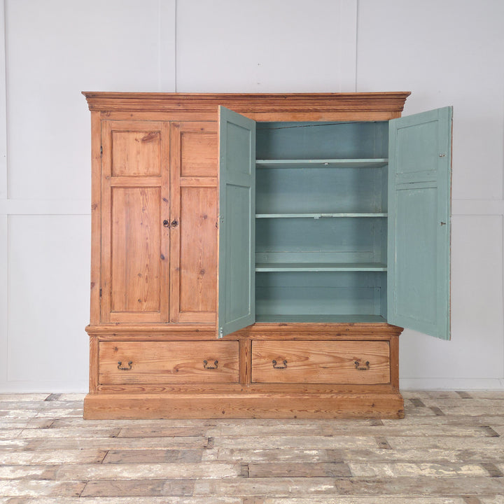 An antique Victorian pine housekeeper’s cupboard dating from the 19th century, with twin panelled doors to the upper section, fixed internal shelving, two base drawers with brass handles, and a simple plinth base.