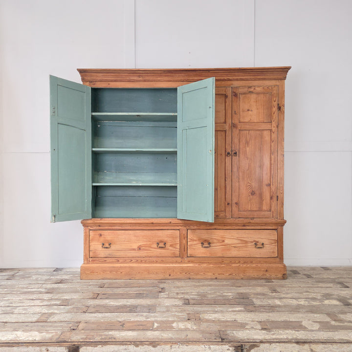 The antique English pine housekeeper’s cupboard has a tall upper section with double panelled doors opening to blue-painted shelves, set above two large base drawers, all raised on a plinth base with a cornice top.