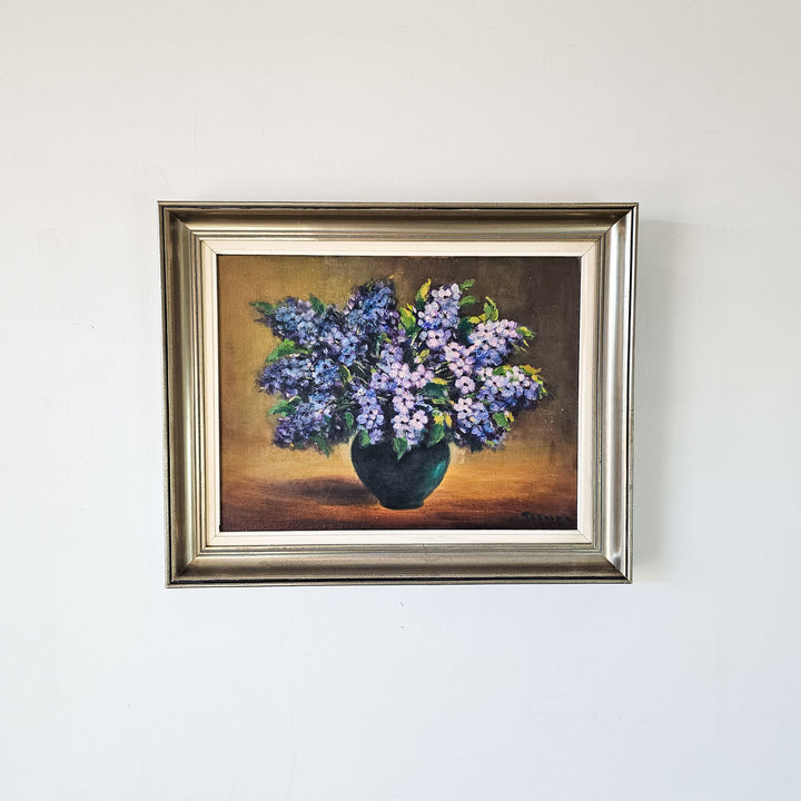 A frontal view of a vintage oil painting on board, featuring a blooming bouquet of purple flowers in a vase.