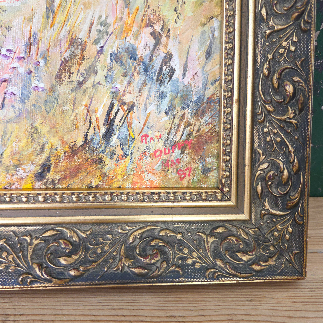An oil on board vintage landscape painting housed in a decorative gilt frame. The composition shows a rural country house scene with outbuildings, meadow grasses in warm yellows and oranges, and rolling hills in deep greens. A naïve style work that captures the charm of the British countryside.