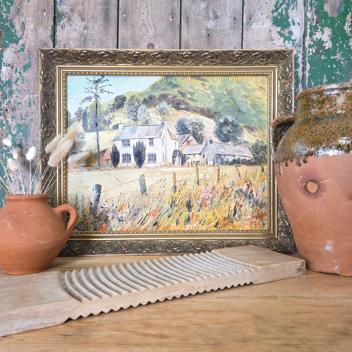 A vintage oil on board landscape painting featuring a rustic countryside scene with a large country house and outbuildings in a meadow, framed by rolling hills. The work is presented in a decorative gilt frame, combining warm golden yellows, oranges and deep green tones for an atmospheric rural view.