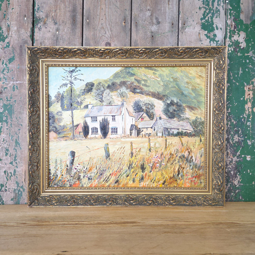 Vintage 20th century oil on board landscape painting showing a country house with surrounding outbuildings, nestled in a meadow. Rolling hills and green trees form the backdrop, with the whole piece displayed in an ornate gilt frame. The colour palette blends earthy yellows, oranges and moody greens.