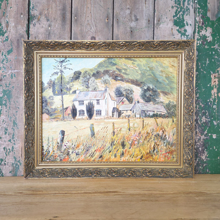 Vintage 20th century oil on board landscape painting showing a country house with surrounding outbuildings, nestled in a meadow. Rolling hills and green trees form the backdrop, with the whole piece displayed in an ornate gilt frame. The colour palette blends earthy yellows, oranges and moody greens.
