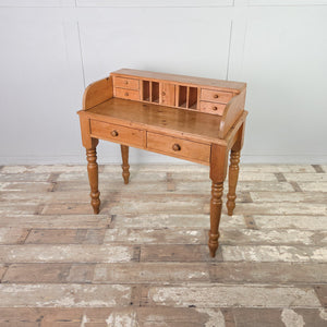 A vintage pine desk raised on four turned legs with two front drawers. The farmhouse style gallery top holds pigeonholes, four small drawers and a miniature cupboard, giving the piece an organised upper layout.