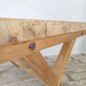 A farmhouse style vintage pine table featuring an X-frame trestle base and substantial rectangular top. Stripped back and wax finished, the table shows natural pine grain. Practical and versatile, it could serve as a rustic dining table, home office desk, or hallway console for everyday us