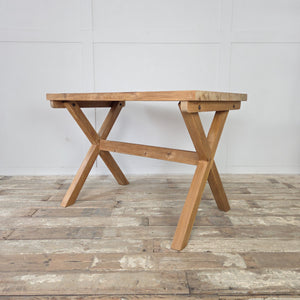 A rustic vintage pine table with trestle construction and an X-frame base. The thick pine top has a stripped finish sealed with wax, showing natural variations in the grain. This farmhouse style piece is highly versatile, used as a kitchen dining table, a study desk, or a hallway console.