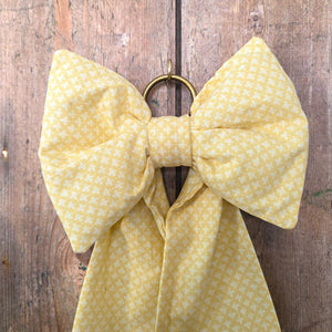 A handmade vintage Country House Bow crafted in yellow patterned fabric. Each bow is structured with a padded form, elongated tails, and finished with an antiqued brass hanging ring, created in limited batches for unique decorative use.