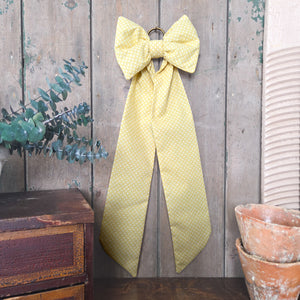Handmade decorative bow created from vintage yellow patterned fabric, with a padded body and tapering tails. This Jennings & Cox Country House Bow is completed with a brass hanging ring and styled for rustic interiors or weddings.