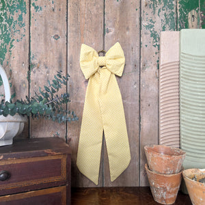 A decorative handmade bow crafted from yellow vintage patterned fabric, padded and shaped with elongated tails. Finished with a brass hanging ring, this Country House Bow is versatile for use on doors, mantels, or as a thoughtful gift.