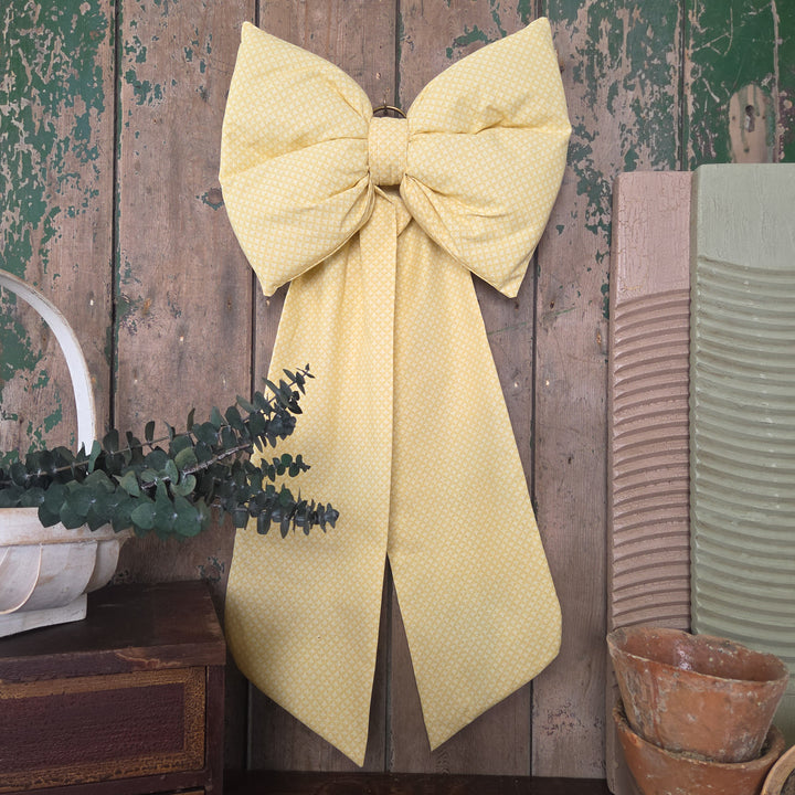 A handmade decorative bow created from genuine vintage fabric in a yellow patterned design. Part of the Jennings & Cox Country House collection, this bow has a padded form, elongated tails, and brass hanging ring for versatile interior styling.