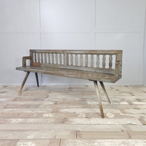 An antique Romanian pine bench with original grey distressed paint, decorative cut-out slatted back, and splayed stick legs
